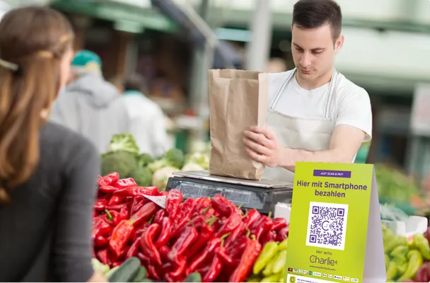 Ein Mann in einem Wochenmarktstand wiegt Gemüse in einer Papiertüte ab. Vor ihm steht ein Qr-Code von Pay with ME.