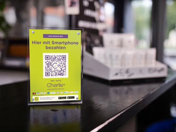 <p>Aufsteller mit einem QR-Code von Pay with ME steht auf einem schwarzen Tresen.</p>